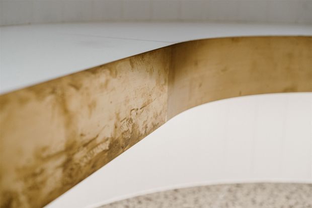 Close-up architectural detail of a curved, layered staircase or sculptural built form. A warm mottled timber or wood-effect vertical band curves between smooth white surfaces, with a speckled floor visible at the bottom. The image is abstract and does not clearly establish a specific room.