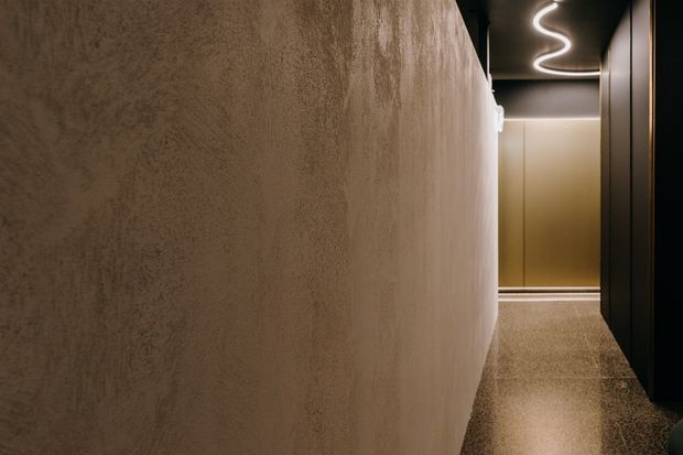 A narrow, dimly lit contemporary hallway or entry corridor with a textured warm taupe plaster wall dominating the left side, dark built-in wall panels on the right, and a polished speckled stone or terrazzo floor. A sculptural looping linear ceiling light provides a soft glow, while warm beige cabinetry or wall paneling is visible at the end of the corridor.