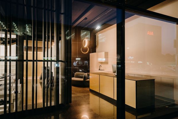 A dark, sophisticated commercial interior viewed through black-framed glass and vertical metal screens. The space features a compact reception or display counter with pale wood cabinetry and a white benchtop, polished dark flooring, exposed dark ceiling elements, and warm accent lighting. Reflections and illuminated signage are visible in the glazing, creating a polished showroom or office atmosphere.