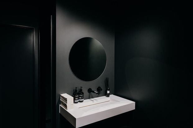A moody, minimalist bathroom vanity in a predominantly black interior. A floating white rectangular basin with integrated benchtop and wall-mounted black tap sits beneath a round frameless mirror. Black wall finishes, a dark door, restrained toiletries, and subtle lighting create a high-contrast, refined atmosphere.