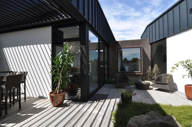 A bright contemporary courtyard terrace connecting several dark-clad building volumes. The space features grey timber decking, black-framed glazing, a slatted pergola casting strong linear shadows, outdoor dining furniture, lounge chairs, potted plants, ornamental grasses, and a small landscaped garden area with rocks. The architecture has crisp rectilinear forms and a restrained charcoal, grey, and white palette.