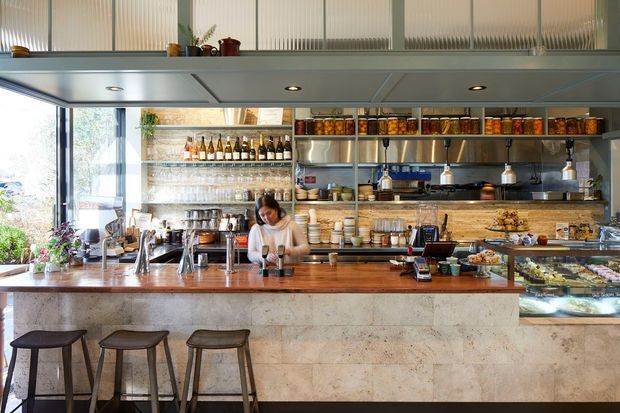 Bright contemporary café interior viewed from the customer side, with a long stone-clad counter and timber benchtop, bar stools, extensive open shelving, hanging pendant lights, display cases, and a staff member preparing drinks. Large windows bring in natural light, while the overhead structure and fluted translucent glazing add architectural character.