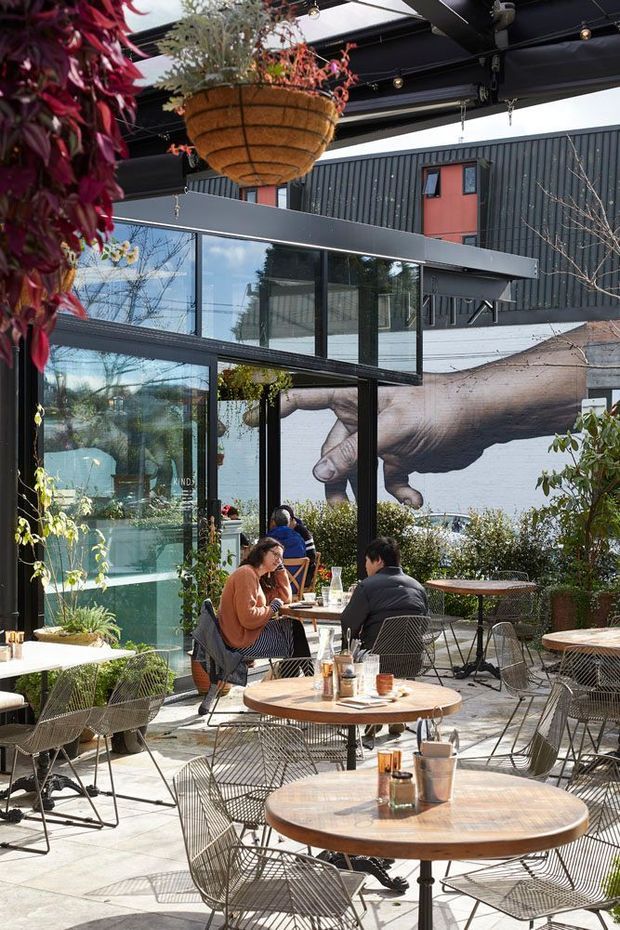 A bright urban outdoor café courtyard or terrace with numerous round wooden tables and wire-frame chairs. The space is enclosed by extensive black-framed glazing and a dark corrugated metal upper facade, with overhead beams, string lighting, and hanging planters. A large mural of a hand reaching toward another hand forms a prominent backdrop, while trees and planting create a leafy atmosphere around the seating area.
