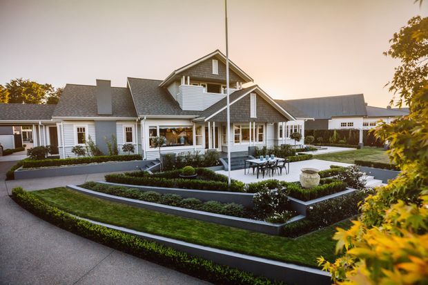 A large, multi-wing suburban residence with a pitched gable roof, white weatherboard-style exterior, dark grey roofing, covered verandas, and prominent white-framed windows. The home is surrounded by formal tiered garden beds, clipped hedges, paved paths, and a central outdoor dining area, viewed from the landscaped front approach in warm evening light.