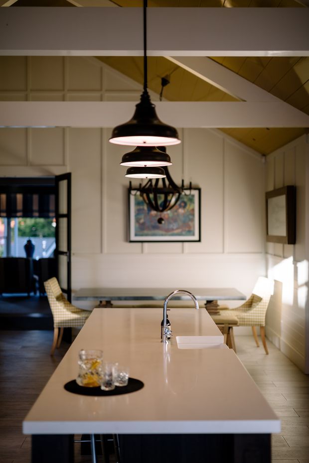 A refined open-plan kitchen and dining area viewed from the kitchen island. The space features a long pale stone island with an integrated sink and curved chrome faucet, dark lower cabinetry, white paneled walls, exposed white structural beams, and a warm timber vaulted ceiling. Black pendant lights hang above the island, while a chandelier, framed artwork, upholstered dining chairs, and a dining table occupy the background. Strong daylight enters from the right and through an open glazed doorway on the left.