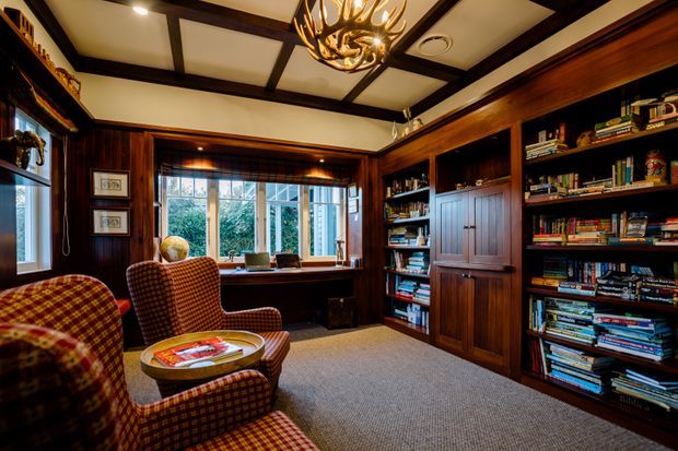 A richly detailed traditional library or home office with extensive dark timber built-in bookshelves and cabinetry, a built-in desk beneath a large window, patterned upholstered armchairs, carpeted flooring, and a decorative antler-style chandelier. The room has warm, moody lighting and strong wood panelling with coffered ceiling beams.