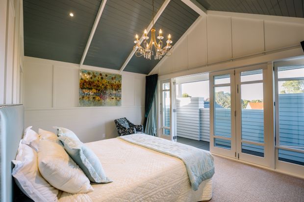 Bright, refined bedroom with a large upholstered bed dressed in white and pale blue bedding. The room features white painted wall panelling, a dark blue-grey vaulted plank ceiling with exposed white beams, a crystal chandelier, carpeted flooring, and extensive glazed doors opening toward an outdoor balcony or terrace. Coastal light and layered traditional detailing create an elegant Hamptons-inspired atmosphere.