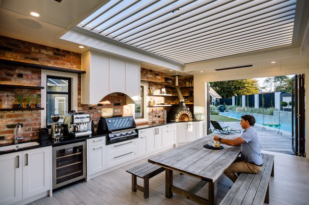 A spacious indoor-outdoor entertaining area combining a white built-in kitchen with a brick feature wall, timber shelving, barbecue and pizza oven, and a large rustic dining table with bench seating. A retractable louvred skylight brings daylight into the room, which opens directly onto a landscaped poolside terrace.