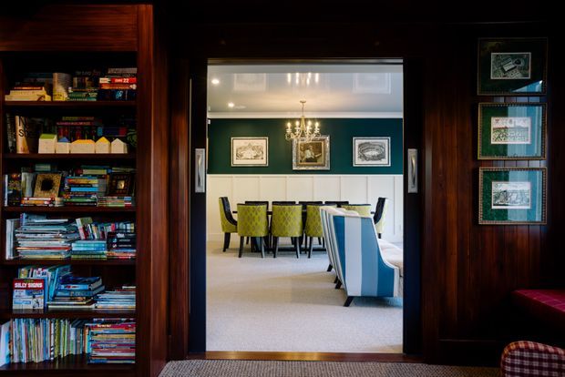 A richly detailed dark timber-lined interior viewed through a wide doorway into a formal dining room. Built-in bookshelves filled with books occupy the left side, while framed artwork lines the right wall. The adjoining dining room has deep teal walls, white wainscoting, a central chandelier, a long dining table with bright patterned chairs, and upholstered blue-and-white seating. The overall composition feels layered, eclectic, and traditionally styled.