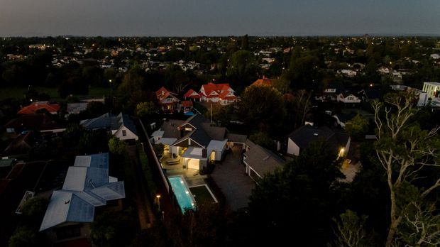 Aerial dusk view over a suburban residential neighbourhood, centred on a modern detached home with a narrow illuminated swimming pool, surrounding paved outdoor areas, pitched roofs, and mature trees. The broader suburb extends across the background under a dark evening sky.