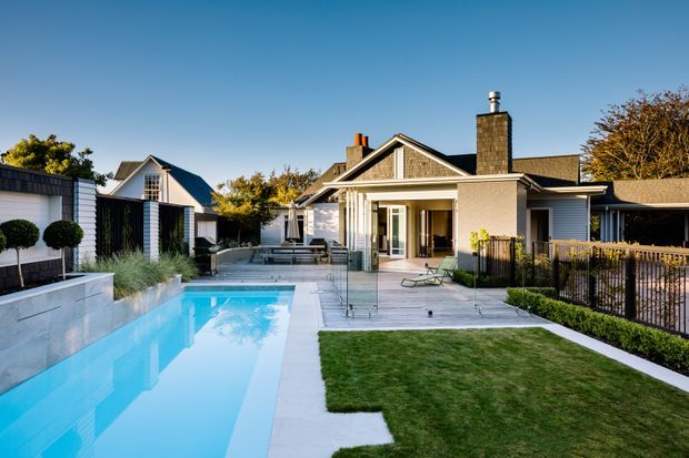 A landscaped residential backyard with a long rectangular swimming pool, paved pool terrace, manicured lawn, timber-and-painted exterior buildings, gabled roofs, covered outdoor entertaining area, and a prominent stone-clad chimney. The scene is bright and contemporary with clean-lined fencing, planting, and outdoor seating.