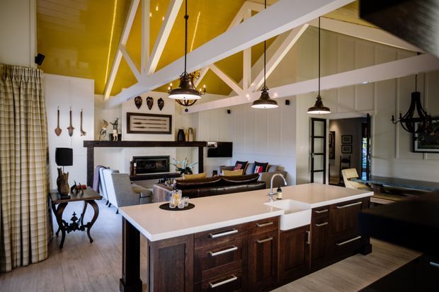 Open-plan kitchen and living area with a large dark timber island, pale benchtops, pendant lighting, and a lounge beyond. The room features a dramatic yellow vaulted ceiling with exposed white structural beams, white vertical wall panelling, dark cabinetry, and a mix of traditional and contemporary furnishings.