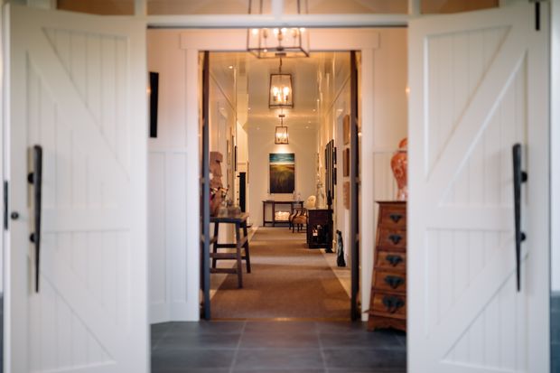 View through open white barn-style double doors into a long, warm-toned interior hallway. The entry has dark large-format tiled flooring, white walls and trim, exposed timber ceiling beams, and multiple hanging lantern-style lights aligned down the corridor. A console table, chest of drawers, framed artwork, and decorative furnishings line the hall, creating a welcoming transitional entry sequence.