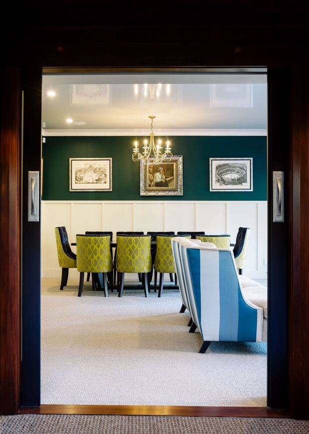 A refined dining room viewed through dark timber double doors, featuring a long dining table with lime-green patterned upholstered chairs, a blue-and-white striped armchair in the foreground, pale carpet, white wall panelling, and a deep green upper wall. A crystal chandelier hangs above the table, with framed artwork arranged behind it. The room has a formal, richly coloured and traditional character.