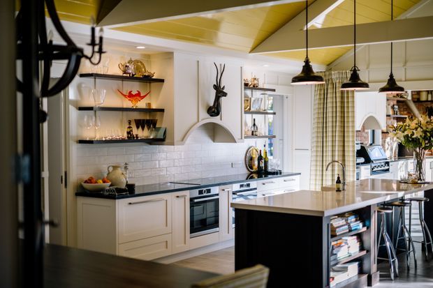 A refined traditional kitchen with white panelled cabinetry, a dark stone island benchtop, white subway-tile splashback, integrated cooking appliances, open display shelving, and a breakfast bar with pendant lights. A glossy mustard-yellow ceiling adds a distinctive feature accent, while a glazed door and adjoining dining area are visible to the right.