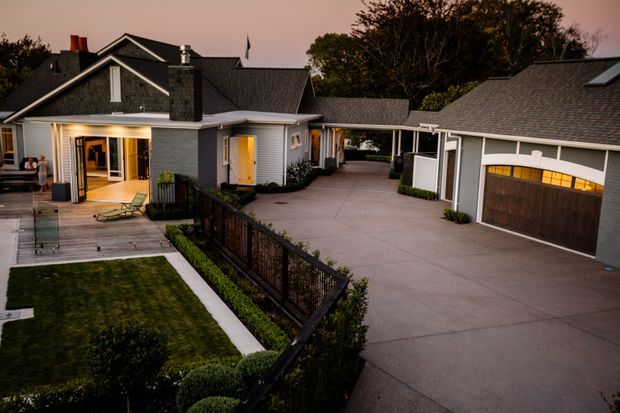 Dusk exterior view of a substantial residential property with a gabled main house, covered entry and porch, landscaped front lawn, paved driveway, and a separate detached garage. The home features grey shingle-style roofing, light horizontal cladding, dark timber accents, warm-lit windows, and a connecting covered passage between buildings.