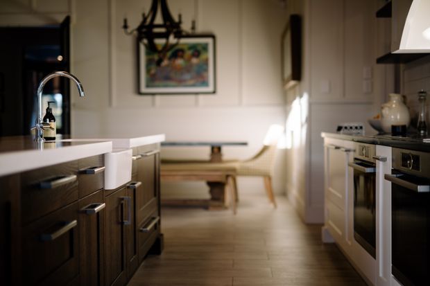 Warm, traditional-style kitchen viewed along a central galley layout, with dark timber cabinetry and a white island benchtop in the foreground. The kitchen includes stainless steel appliances, a tall curved chrome tap, open shelving, and a softly lit dining or sitting nook beyond with paneled walls and a framed artwork.