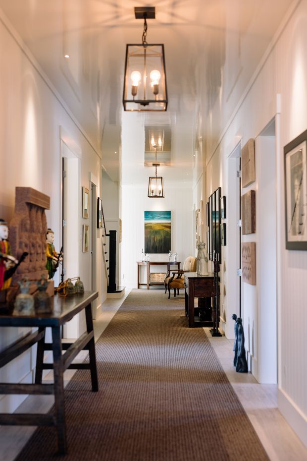 A long, narrow residential hallway with white paneled walls, a glossy white ceiling, pale timber flooring, and a broad textured runner. Black-framed pendant lanterns hang along the corridor, while console tables, framed artwork, decorative objects, and a distant desk-style setting line the walls. The space has a refined, gallery-like traditional feel with warm ambient lighting.