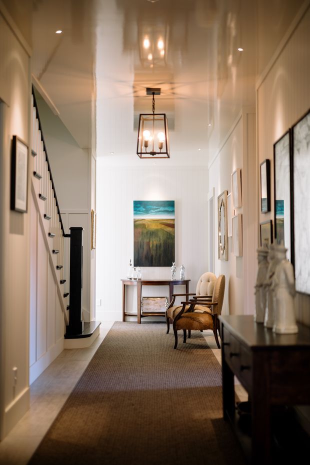 A refined residential hallway and entry corridor with white panelled walls, a glossy white ceiling, built-in stair storage, framed artwork, a dark console, and a narrow console table at the far end. Two upholstered vintage-style chairs sit beside the table, while a warm lantern pendant and recessed lighting create an elegant, inviting atmosphere. A runner extends through the corridor.