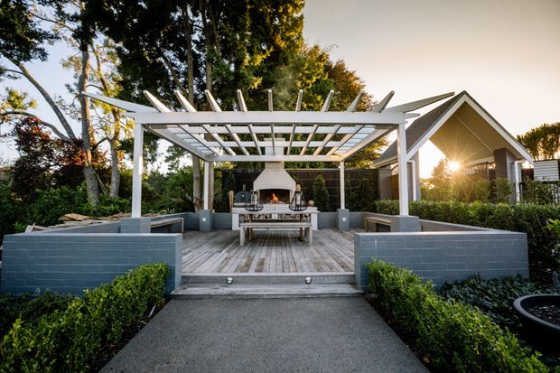 A landscaped outdoor entertaining area featuring a white timber pergola with open slatted roof, built-in seating walls, a central outdoor fireplace and dining table, and a timber deck reached by a paved path. A gabled house is visible behind the garden.