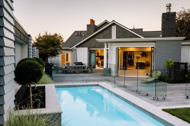 Exterior view of a contemporary grey-clad house surrounding a landscaped courtyard and rectangular swimming pool. The home combines gabled roof forms, dark shingle-like cladding, white horizontal weatherboards, large glazed openings, and a covered outdoor entertaining area. Timber decking, frameless glass pool fencing, clipped shrubs, and warm interior lighting create a refined residential setting.