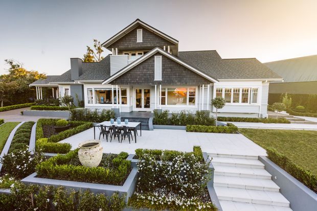 Exterior view of a large, symmetrical suburban house with white weatherboard cladding, grey shingle-style gabled roofs, prominent dormer-like gable forms, and white-trimmed windows. The property features a formal landscaped garden with geometric hedges, paved terraces, outdoor dining furniture, and broad steps leading to the home.