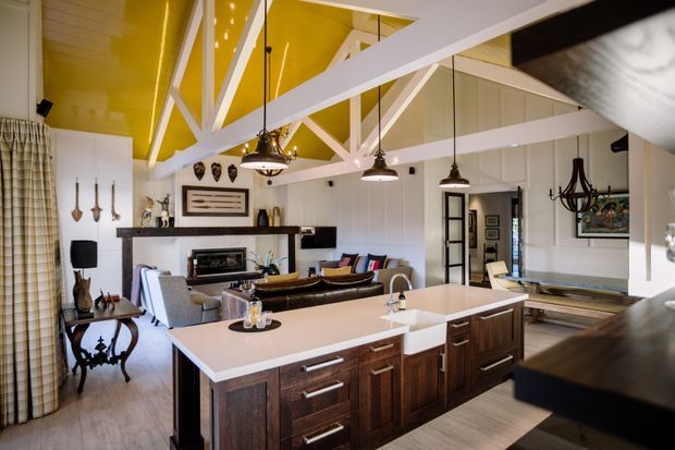 Open-plan kitchen and living area with a large dark timber island, pale benchtops, white cabinetry and exposed white roof trusses. The room features a striking glossy mustard-yellow ceiling, pendant lighting, a fireplace, and an adjacent lounge with dark leather seating.