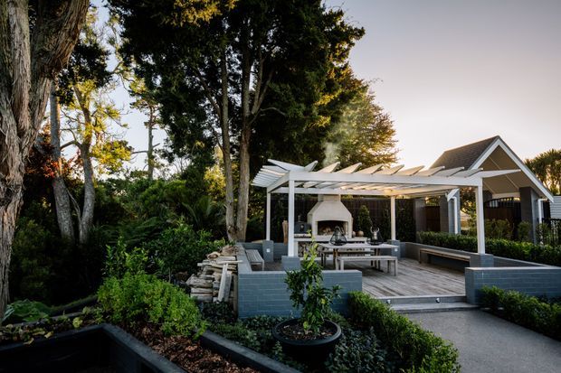 A landscaped outdoor entertaining area beside a gabled house, featuring a raised timber deck, large white pergola with retractable-looking shade panels, built-in seating, an outdoor fireplace, and a dining table. Dense mature planting and trees frame the space, with dark painted masonry retaining walls and a paved path leading to the deck.