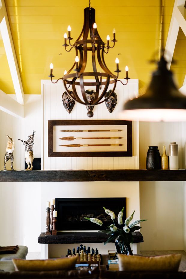 A richly styled interior centered on a dramatic dark timber fireplace and mantel, with a vivid yellow painted timber-lined ceiling, white structural beams and wall panelling. A large dark metal multi-arm chandelier hangs above framed spear artwork, sculptural decor, and a dining table with upholstered chairs in the foreground. The room has a bold, eclectic lodge-inspired character.