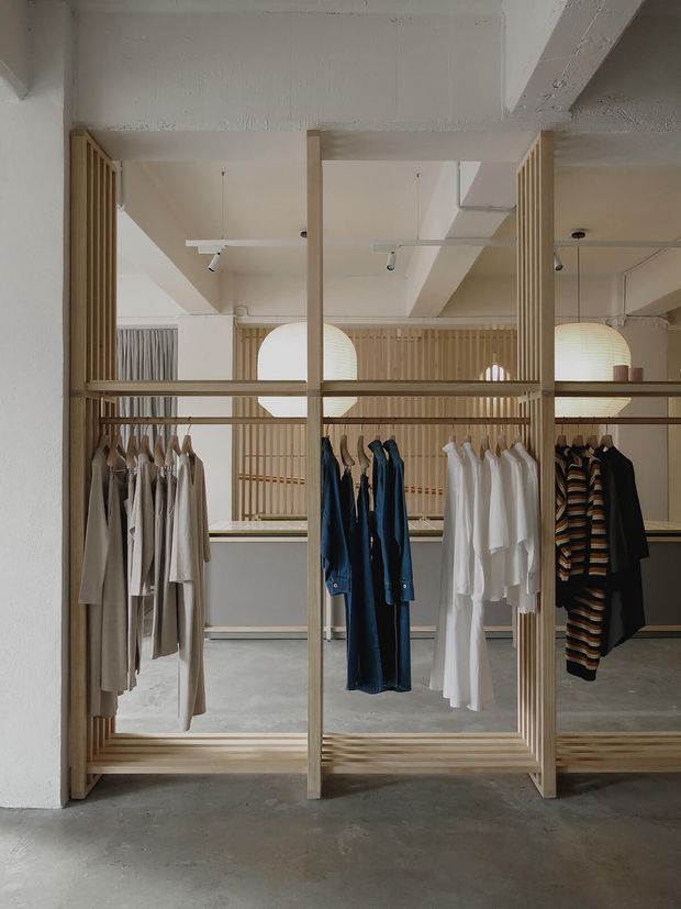 A minimalist clothing retail interior with open, floor-to-ceiling timber slat garment displays and hanging rails. Neutral clothing is arranged across several bays, with pale timber screens, large spherical pendant lights, track lighting, exposed white ceiling elements, and a polished grey concrete floor creating a calm, refined boutique atmosphere.