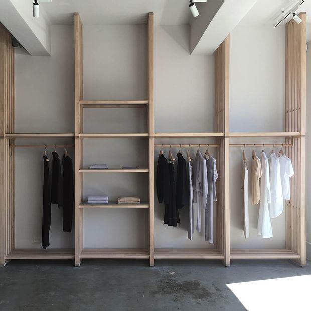 A minimalist walk-in wardrobe with open, floor-to-ceiling timber-framed shelving and hanging rails. Neutral grey walls, exposed concrete flooring, and white ceiling beams create a clean, restrained interior. Clothes are arranged by dark, grey, and light tones, with strong natural light entering from the right.