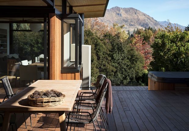 A contemporary timber-clad home opens onto a spacious outdoor timber deck with a dining table and woven chairs. Large glazed doors connect the deck to the interior, while a dark built-in spa or plunge pool structure sits at the far right. Dense landscaping and a rugged mountain backdrop create a strong alpine setting. The image is brightly lit with warm natural timber, dark frames, and autumn foliage.