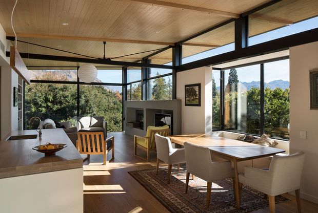 A bright open-plan dining and living area with floor-to-ceiling glazing overlooking trees and distant mountains. The room features a timber-lined ceiling, dark exposed structural beams and window frames, warm timber flooring, a dining table with upholstered chairs, a compact sitting area, and a central fireplace built into a pale wall. A kitchen island is visible in the foreground.