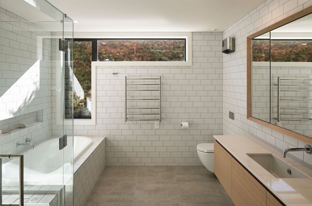 Bright contemporary bathroom with white subway-tiled walls, a freestanding bathtub beside a frameless glass shower enclosure, wall-hung toilet, heated towel rail, and a long timber-veneer vanity with an integrated basin. A large timber-framed mirrored cabinet reflects the vanity and towel rail. Narrow horizontal windows bring in natural light and views of vegetation.