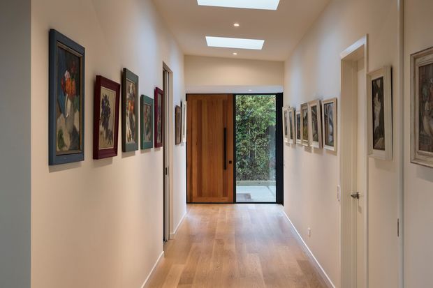 A long, narrow residential hallway lined with framed artwork on both walls. The corridor has warm light timber flooring, smooth off-white walls, white doors, recessed ceiling lights, and rectangular skylights. At the end is a large timber pivot entry door beside full-height glazing opening toward greenery, creating a bright gallery-like passage.