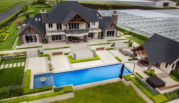 Aerial view of a large, landscaped residential estate with a dark pitched-roof main house, stone and light-rendered facade, multiple gabled sections, extensive terraces and paving, a long swimming pool, a detached pool pavilion, manicured hedges, and a large glasshouse or greenhouse structure in the background.