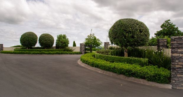 A formal landscaped driveway or entrance with clipped spherical topiary trees, low manicured hedges, ornamental grasses, and dark stacked-stone pillars and walls. The wide asphalt drive curves through the garden beneath an overcast sky, creating a refined, highly maintained estate approach.