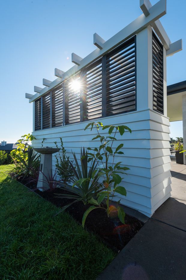 Exterior architectural detail of a contemporary coastal-style home featuring white horizontal weatherboard cladding, dark horizontal louvres set within a projecting screen, and exposed white structural beams. A landscaped garden and paved walkway border the facade under a clear blue sky.