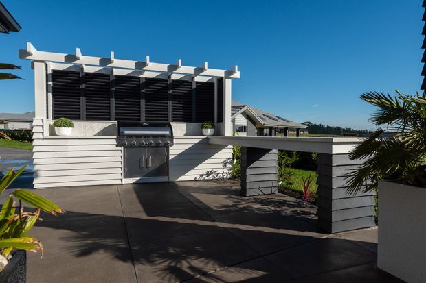 A contemporary outdoor entertaining area with a built-in stainless-steel barbecue beneath a white pergola and dark horizontal privacy screens. The space features a broad dark paved deck, raised planter walls, integrated bench-like structures, and tropical planting, with neighbouring pitched-roof homes visible beyond.