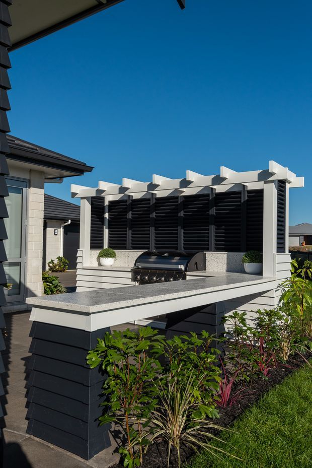 A contemporary outdoor entertaining area beside a house, featuring a built-in barbecue set into a raised, speckled stone-look benchtop with integrated seating or serving ledges. The structure has dark horizontal cladding, white posts and pergola-like overhead beams, with black louvred screening behind the grill. Garden planting surrounds the elevated platform, and neighbouring modern homes are visible under a clear blue sky.