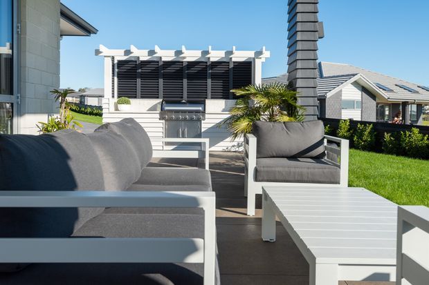 Bright contemporary outdoor lounge area with white-framed grey upholstered seating and a slatted coffee table on a paved terrace. A white pergola-like structure with dark horizontal louvres, outdoor fireplace or feature wall, potted palms, clipped hedges, and adjacent modern homes are visible in the background.