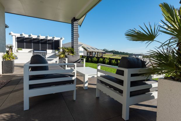 A bright, modern outdoor terrace overlooking a landscaped suburban setting. The covered patio features two white slatted lounge chairs with dark cushions, a matching low coffee table, large planters, palms, and a contemporary pergola-like screen structure. The space has a clean, open feel with views toward neighbouring rooftops and greenery.