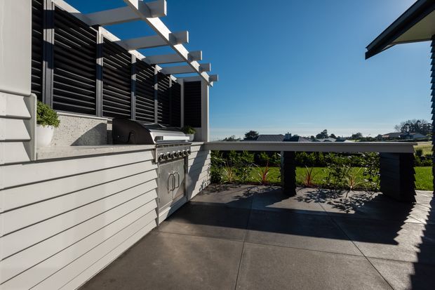 A contemporary rooftop or elevated outdoor entertaining area with a built-in stainless-steel barbecue, pale horizontal-clad cabinetry, dark louvred privacy screening, and a white pergola structure. The paved terrace overlooks a suburban landscape and is edged by a dark balustrade with integrated planting.