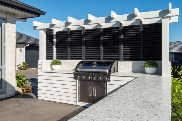 A bright contemporary outdoor kitchen and barbecue area featuring a large stainless-steel grill, pale speckled stone benchtops, white horizontal cladding, and a white pergola structure with black louvred privacy screens. Potted plants add greenery, with neighbouring rendered buildings visible around the paved courtyard.