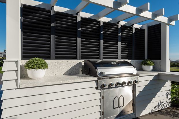 A bright contemporary outdoor kitchen featuring a stainless-steel built-in gas barbecue set into a white horizontal-clad counter beneath a white pergola. Black louvred privacy screens line the rear wall, with concrete-look benchtops and potted greenery adding contrast.