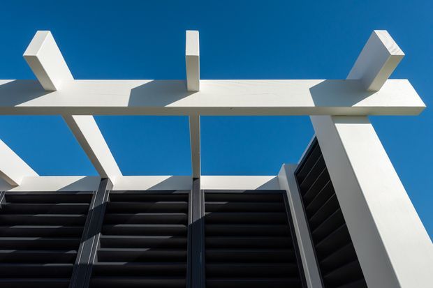 Upward-looking architectural detail showing a bold white structural pergola or roof frame with projecting angled beams, dark horizontal louvres below, and strong geometric shadows against a clear blue sky.