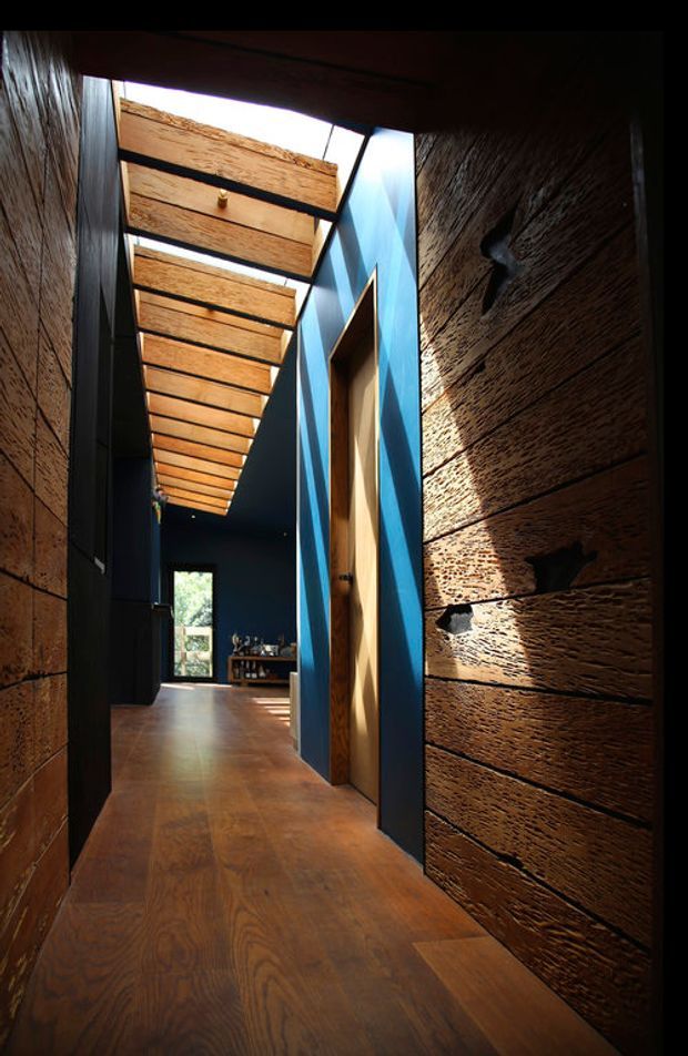 A narrow, dramatic interior hallway lined with dark textured timber wall cladding and warm timber flooring. Deep blue painted walls frame a timber doorway, while a series of exposed timber slats overhead creates a partially open skylight or clerestory effect, casting strong sunlight and shadows into the corridor. A living area is visible at the far end.