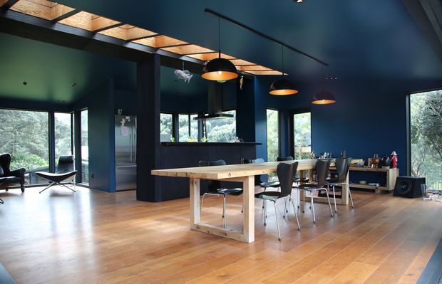 Open-plan dining and living area with a long timber dining table, mid-century style chairs, dark navy-blue walls and ceiling, exposed timber roof glazing/skylight structure, timber flooring, pendant lights, and extensive windows overlooking greenery. A compact kitchen is visible behind the dining area.