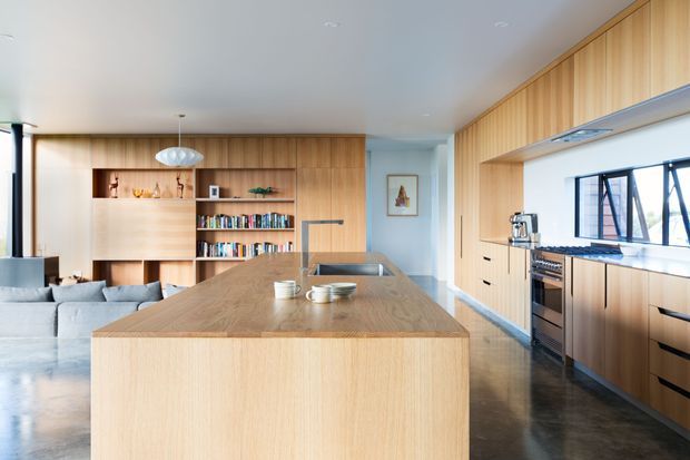 Open-plan contemporary kitchen and living area with extensive light timber cabinetry and wall panelling. A large timber island with integrated sink dominates the foreground, with black appliances, dark polished flooring, open shelving, a built-in bookcase, and a pale grey sectional sofa visible beyond.