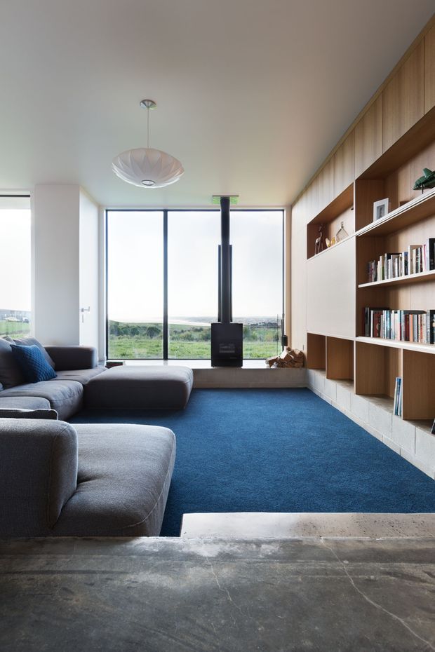 A contemporary living room with a large charcoal sectional sofa, deep blue carpet, and a full-height built-in timber library wall with open shelving and cabinetry. Floor-to-ceiling glazing overlooks a green rural landscape, while a suspended sculptural pendant and central black wood-burning stove add focal points. The foreground has a polished grey concrete floor and a raised window seat.